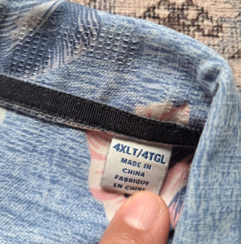 K&ouml;nnte beinhalten: Nahaufnahme eines blauen Hemdes mit floralem Muster. Das Etikett lautet "4XLT/4TGL" und "Made in China". Der Stoff hat eine strukturierte Optik mit einem schwarzen Kragendetail. Das Etikett sagt auch "Fabriqu&eacute; en Chine".