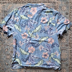 K&ouml;nnte beinhalten: Kurz&auml;rmeliges Hemd mit tropischem Blumenmuster. Das Hemd hat einen hellblauen Hintergrund mit rosa Hibiskusbl&uuml;ten, gr&uuml;nen Bl&auml;ttern und lila Akzenten. Das Hemd ist aus leichtem Stoff gefertigt.