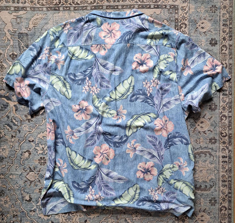 K&ouml;nnte beinhalten: Kurz&auml;rmeliges Hemd mit tropischem Blumenmuster. Das Hemd hat einen hellblauen Hintergrund mit rosa Hibiskusbl&uuml;ten, gr&uuml;nen Bl&auml;ttern und lila Akzenten. Das Hemd ist aus leichtem Stoff gefertigt.