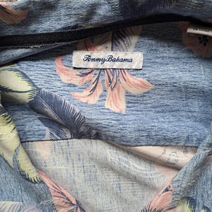 K&ouml;nnte beinhalten: Hellblaues Tommy Bahama-Hemd mit einem Blumenmuster in Rosa und Wei&szlig;. Das Hemd hat einen Kragen und ein schwarzes Etikett mit dem Markennamen. Der Stoff hat ein strukturiertes Aussehen.