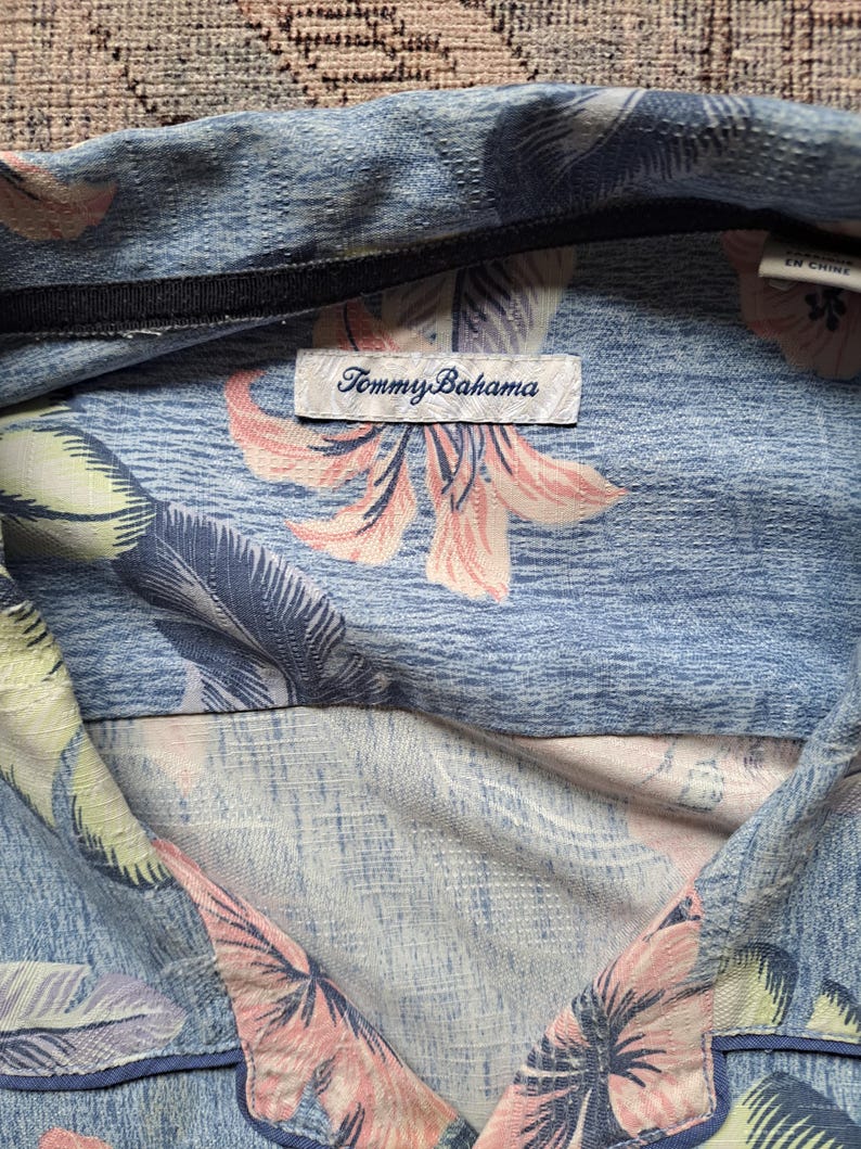K&ouml;nnte beinhalten: Hellblaues Tommy Bahama-Hemd mit einem Blumenmuster in Rosa und Wei&szlig;. Das Hemd hat einen Kragen und ein schwarzes Etikett mit dem Markennamen. Der Stoff hat ein strukturiertes Aussehen.