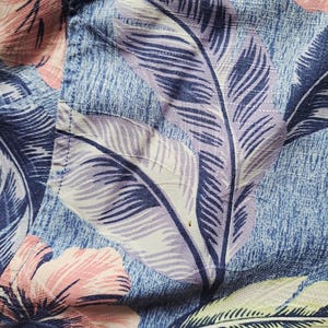 K&ouml;nnte beinhalten: Nahaufnahme eines blauen Stoffes mit floralem Muster. Das Design zeigt gro&szlig;e, stilisierte Bl&auml;tter in Lila-, Wei&szlig;- und Gr&uuml;nt&ouml;nen sowie rosa Hibiskusbl&uuml;ten. Der Stoff hat ein strukturiertes Aussehen, was auf ein gewebtes Material hindeutet.