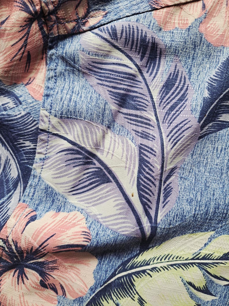 K&ouml;nnte beinhalten: Nahaufnahme eines blauen Stoffes mit floralem Muster. Das Design zeigt gro&szlig;e, stilisierte Bl&auml;tter in Lila-, Wei&szlig;- und Gr&uuml;nt&ouml;nen sowie rosa Hibiskusbl&uuml;ten. Der Stoff hat ein strukturiertes Aussehen, was auf ein gewebtes Material hindeutet.