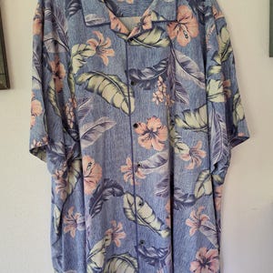 K&ouml;nnte beinhalten: Blaues Button-Down-Hemd mit einem Blumenmuster aus rosa Hibiskusbl&uuml;ten und gro&szlig;en gr&uuml;nen und lila Bl&auml;ttern. Das Hemd ist kurz&auml;rmlig und hat eine lockere Passform, die an einem Holzb&uuml;gel h&auml;ngt.