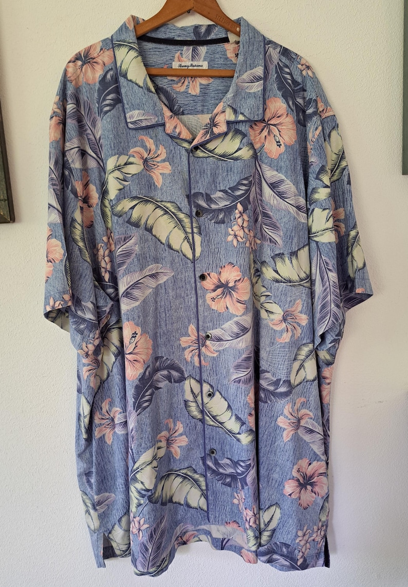 K&ouml;nnte beinhalten: Blaues Button-Down-Hemd mit einem Blumenmuster aus rosa Hibiskusbl&uuml;ten und gro&szlig;en gr&uuml;nen und lila Bl&auml;ttern. Das Hemd ist kurz&auml;rmlig und hat eine lockere Passform, die an einem Holzb&uuml;gel h&auml;ngt.