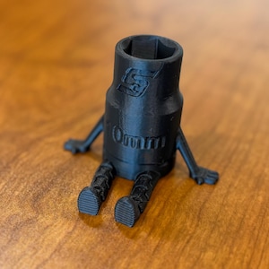 Puede incluir: Una llave de vaso negra impresa en 3D con brazos y piernas, lo que le da una apariencia de dibujos animados. La llave está sentada sobre una superficie de madera. La llave de vaso tiene el número "10mm" impreso.