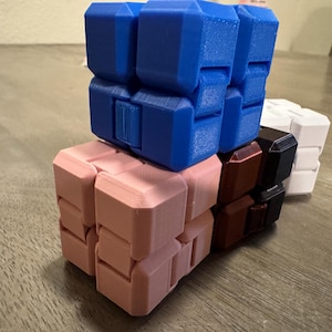 May include: A collection of colorful fidget cubes in blue, pink, brown, black, and white. The cubes are stacked on top of each other, showcasing their geometric design and various color combinations. The cubes are made of plastic.