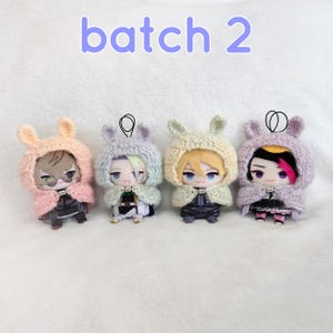 Peut inclure: Quatre petites poupées portant des capuches à oreilles de lapin tricotées dans des tons de rose, de lavande et de crème. Chaque poupée a une coiffure et une tenue uniques. Le texte "batch 2" est affiché en violet au-dessus des poupées.