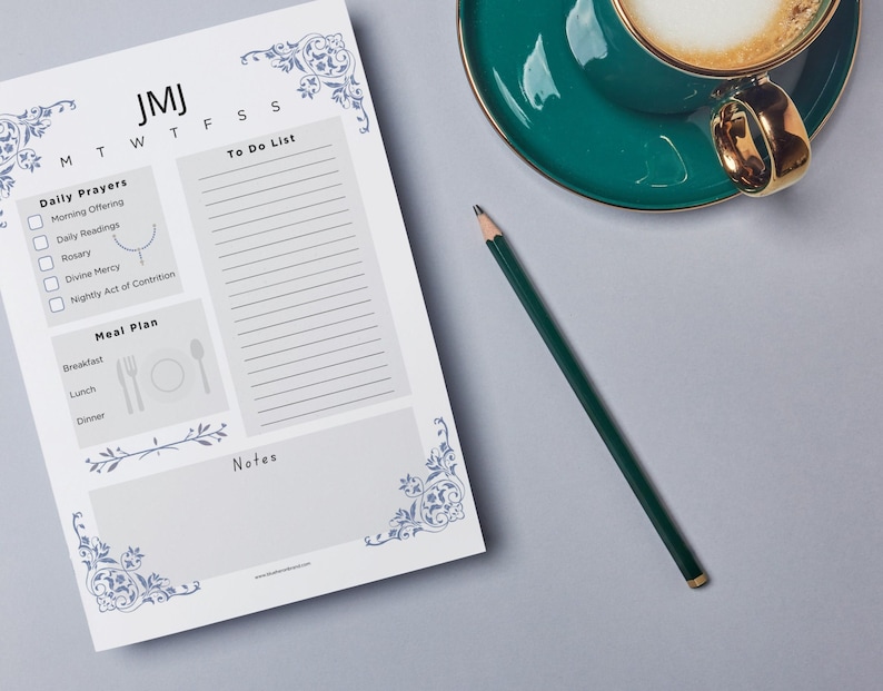 May include: A white printable daily planner with blue floral designs on the corners. The planner has sections for daily prayers, a meal plan, a to-do list, and notes. The planner has the letters JMJ at the top and the days of the week listed below. The planner is on a gray surface with a green cup of coffee and a green pencil.