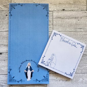 May include: Two blue and white notepad sets with decorative borders. One notepad has a blue background with a drawing of Mary, mother of Jesus, and the text "Hail Mary, Full of Grace, the Lord is with Thee". The other notepad has a white background with a drawing of Mary holding baby Jesus and the text "Blessed is She".