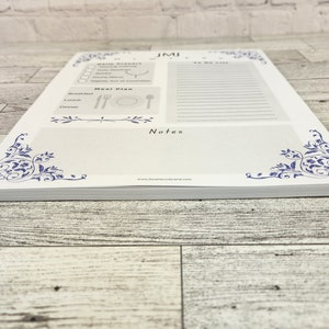 K&ouml;nnte beinhalten: Ein wei&szlig;er Notizblock mit blauem floralem Design in den Ecken und am oberen Rand. Der Notizblock enth&auml;lt Abschnitte f&uuml;r t&auml;gliche Gebete, eine To-Do-Liste, Essensplanung und Notizen. Der Notizblock ist mit vielen Bl&auml;ttern gestapelt.