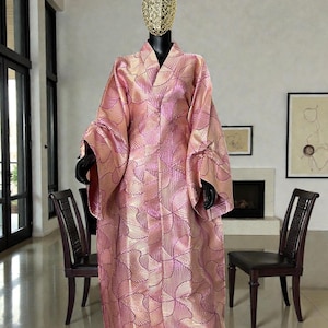 Może przedstawiać: Różowa szata w stylu kimono z wzorem w kształcie wachlarza w ciemniejszym różu. Szata ma szerokie rękawy i dekolt w serek. Szata jest prezentowana na manekinie ze złotą osłoną głowy.