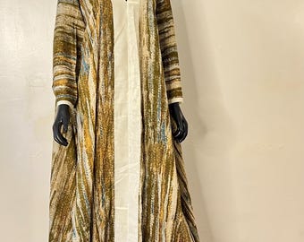 May include: Full-length, patterned dress on a mannequin. The dress features a V-neck, long sleeves, and a cream-colored panel down the front. The fabric has a mix of olive green, brown, and cream colors. A gold head covering is on the mannequin.