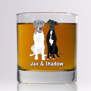May include: A clear glass tumbler with a brown liquid inside. The glass has a black and white illustration of two dogs sitting side by side. The text "Jax & Shadow" is printed below the dogs.
