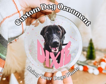 May include: A clear glass round ornament with a black dog photo inside. The dog's name "Dixie" is written in pink letters on the ornament. The ornament has a gold string for hanging. The text "Custom Dog Ornament Using Your Dogs Photo" is written around the ornament.