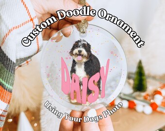 May include: A clear glass ornament with a white background and pink polka dots. A photo of a dog is inside the ornament with the name "Daisy" written in pink letters. The ornament is hanging from a gold string and the text "Custom Doodle Ornament Using Your Dogs Photo" is written around the ornament.