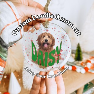 May include: A clear glass Christmas ornament with a red and white snowflake pattern. A brown and white doodle dog is inside the ornament with the name "Daisy" written in green letters. The ornament has a gold string for hanging.