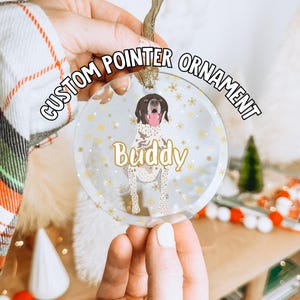 May include: A clear glass Christmas ornament with a gold glitter background and a white German Shorthaired Pointer dog illustration. The dog is wearing a red collar and has a pink tongue sticking out. The ornament has the text "CUSTOM POINTER ORNAMENT" in gold lettering around the top and the name "Buddy" in gold lettering below the dog.