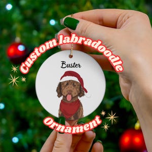 May include: A white ceramic ornament with a brown and white labradoodle wearing a red Santa hat and a red bandana. The ornament has the text "Buster" and "Custom labradoodle ornament" around the edge.