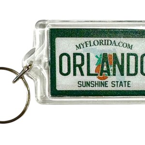 Pode incluir: Chaveiro retangular transparente com um design verde e branco. O design apresenta o texto "MYFLORIDA.COM ORLANDO SUNSHINE STATE". Um anel de metal dourado está preso ao chaveiro.