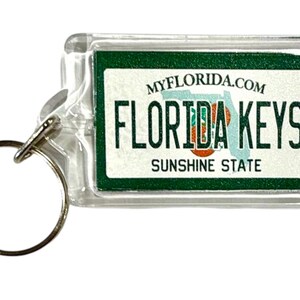 以下が含まれることがあります： シルバーのキーリングが付いた、透明な長方形のキーホルダー。「FLORIDA KEYS」と「SUNSHINE STATE」の文字が入った緑と白のデザイン。ウェブサイト「MYFLORIDA.COM」もキーホルダーに印刷されています。