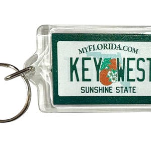 以下が含まれることがあります： 緑色の背景に「KEY WEST」と「SUNSHINE STATE」の文字が入った、透明な長方形のキーホルダー。「MYFLORIDA.COM」の文字と花と果物のグラフィックも含まれています。
