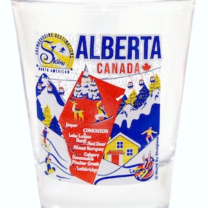 Pode incluir: Um copo de shot de vidro transparente com um design colorido que apresenta o texto "ALBERTA CANADA" e um mapa da província com vários resorts de esqui listados. O design inclui uma cena de montanha vermelha e azul com esquiadores e snowboarders.