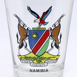 May include: A clear glass shot glass with a colorful image of the Namibian coat of arms. The coat of arms features two oryx, an eagle, and a shield with the Namibian flag. The text "UNITY LIBERTY JUSTICE NAMIBIA" is printed below the coat of arms.