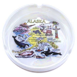 May include: White ceramic ashtray with a colorful map of Alaska. The map features various cities and landmarks, including Anchorage, Fairbanks, Juneau, and Sitka. The ashtray has a raised rim and a smooth surface.