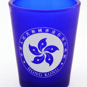May include: A blue shot glass with a white Hong Kong emblem featuring a stylized bauhinia flower with five stars. The text "HONG KONG" is printed below the emblem.