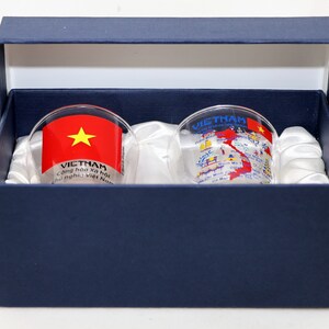May include: Two clear glass shot glasses with a blue box. One glass has a red and yellow Vietnamese flag with the text "VIETNAM Cong hoa xa hoi chu nghia Viet Nam". The other glass has a map of Vietnam with cities and landmarks printed on it.