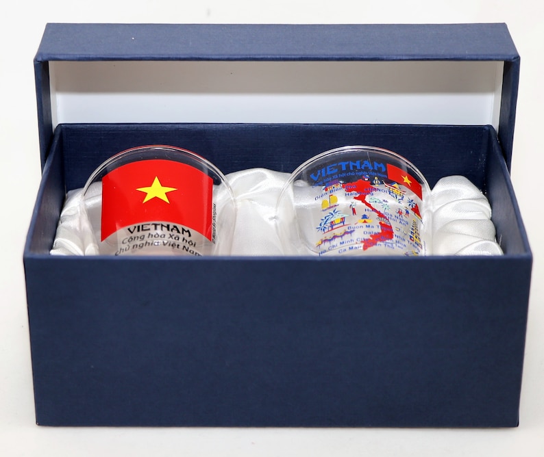May include: Two clear glass shot glasses with a blue box. One glass has a red and yellow Vietnamese flag with the text "VIETNAM Cong hoa xa hoi chu nghia Viet Nam". The other glass has a map of Vietnam with cities and landmarks printed on it.