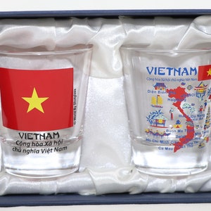 May include: Two clear glass shot glasses with a design featuring the flag of Vietnam and a map of Vietnam with city names. The glasses are in a blue and white box.