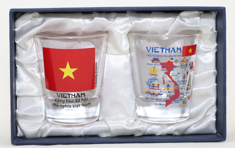 May include: Two clear glass shot glasses with a design featuring the flag of Vietnam and a map of Vietnam with city names. The glasses are in a blue and white box.