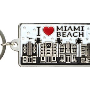 May include: Silver keychain with a white background and a red heart that says "I love Miami Beach". The keychain features a black and white silhouette of a city skyline with palm trees and a crescent moon.