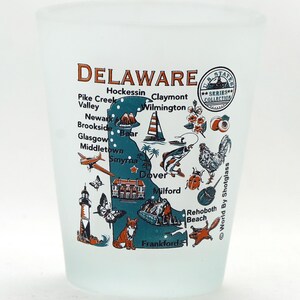 May include: A frosted glass shot glass with a map of Delaware and various illustrations of the state's landmarks, animals, and cities. The text "DELAWARE" is printed in orange at the top of the glass. The text "U.S. STATES SERIES COLLECTION" is printed in a circle at the top right of the glass. The text "World By Shotglass" is printed on the right side of the glass.