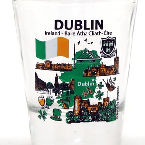 May include: A clear glass shot glass with a green, white, and orange Irish flag, a green shamrock, and a graphic of Dublin, Ireland. The text "Dublin, Ireland - Baile Atha Cliath - Éire" is printed on the glass.