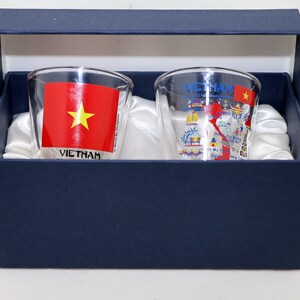 May include: Two clear glass shot glasses with a blue and white design featuring a map of Vietnam and the Vietnamese flag. The glasses are in a blue box with white satin lining.