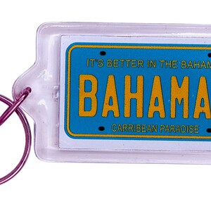 May include: A rectangular keychain with a clear plastic casing. The keychain features a blue license plate design with the word "BAHAMAS" in yellow, and the text "IT'S BETTER IN THE BAHAMAS" and "CARRIBEAN PARADISE". A purple key ring is attached.