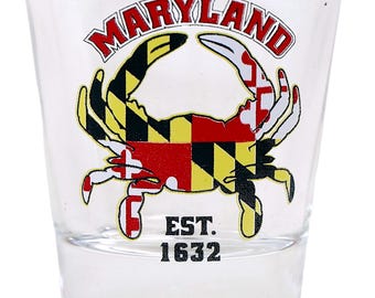 May include: A clear glass shot glass featuring a Maryland crab design. The crab is outlined in white and filled with the Maryland state flag colours of black, yellow, and red. The text "MARYLAND" is printed in red and black above the crab, and "EST. 1632" is printed in black below the crab.