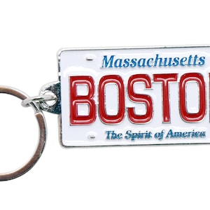 May include: A Boston, Massachusetts license plate keychain. The keychain features a white background with red lettering that spells out "BOSTON". The text "Massachusetts" is above the word "BOSTON" in blue. The phrase "The Spirit of America" is below the word "BOSTON".