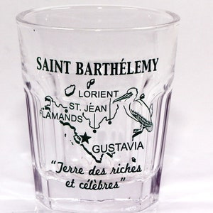 May include: A clear glass shot glass with a map of Saint Barthélemy printed on the side. The map shows the locations of Gustavia, Lorient, St. Jean, and Flamands. The text "Terre des riches et célèbres" is printed below the map.