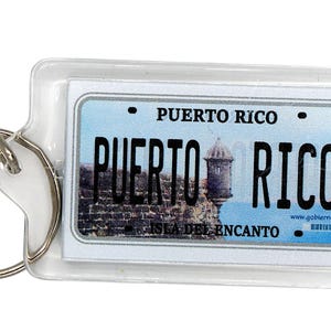 Könnte beinhalten: Ein rechteckiger Schlüsselanhänger mit einem Design eines Nummernschildes von Puerto Rico. Das Schild trägt die Aufschrift "Puerto Rico" und ein Bild einer Festung. Der Schlüsselanhänger enthält einen silbernen Schlüsselring.