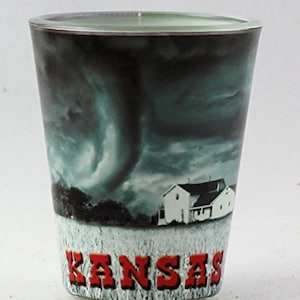 May include: A clear glass shot glass with a printed image of a tornado and a farmhouse with the word "Kansas" in red letters at the bottom.