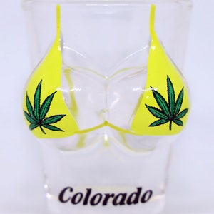 May include: A clear glass shot glass with a yellow bikini design featuring a green marijuana leaf on each side. The word "Colorado" is printed on the bottom of the glass.