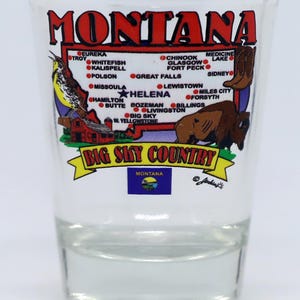 May include: A clear glass shot glass with a colorful illustration of the state of Montana. The illustration includes the state's outline, major cities, and the text "Big Sky Country". The state flag is also included at the bottom of the glass.