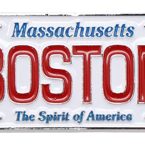 May include: A white metal license plate with blue and red lettering. The plate reads "Massachusetts" and "BOSTON" in large red letters. The bottom of the plate reads "The Spirit of America".