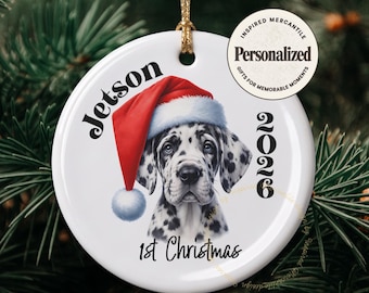 May include: A round white ceramic ornament featuring a Dalmatian puppy wearing a red and white Santa hat. The text reads Jetson, 2026, and 1st Christmas. A gold string is attached to the top for hanging. A circular logo sticker reads Personalized.