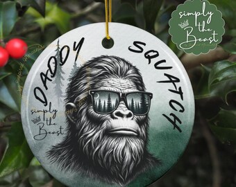 May include: A round white ceramic ornament with a black and white illustration of a Bigfoot wearing sunglasses. The text "Daddy Squatch" is written in a curved font around the Bigfoot. The text "simply the Beast" is written in a smaller font below the Bigfoot.