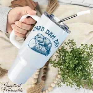 May include: White tumbler with a handle and a silver straw. The tumbler features a blue illustration of a beaver on a log with the text "HARD DAM DAY". The tumbler has a white lid and a white handle.